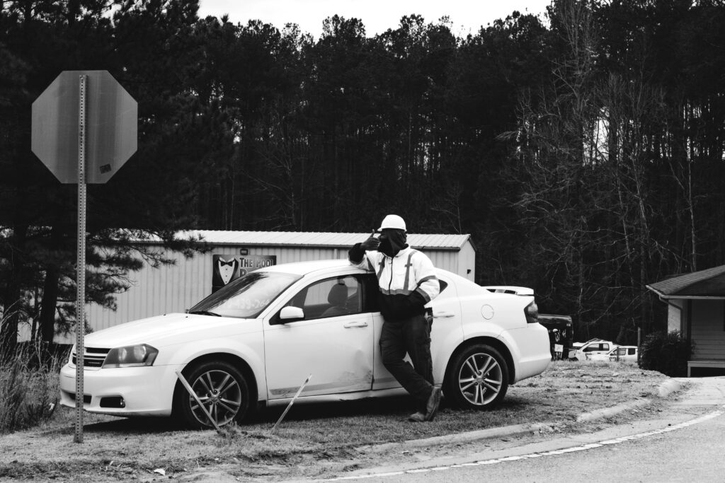 Man standing beside a damaged white car on the roadside calling for help after a minor car accident